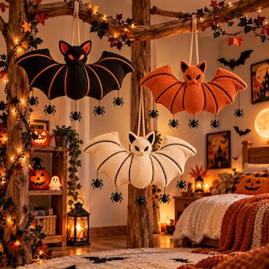 Pode incluir: Três decorações de morcego de pelúcia penduradas num galho de madeira. Um morcego é preto com detalhes laranja, um é laranja sólido e um é creme. Cada morcego apresenta orelhas pontiagudas, detalhes de asas costuradas e pequenas aranhas.