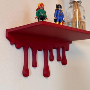 Puede incluir: Una estantería de pared de color rojo mate con un diseño decorativo de goteo en el borde frontal. Dos pequeñas figuras de superhéroes se encuentran sobre la superficie plana junto a un frasco de vidrio transparente con varillas difusoras de madera.