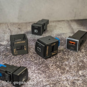 Könnte beinhalten: Eine Sammlung schwarzer elektronischer Komponenten, darunter USB-Ladegeräte und Steuerschalter. Einige Komponenten tragen Aufschriften wie "DRIVER", "UP" und "Ver.7". Eine Komponente zeigt ein Batteriesymbol. Die Komponenten sind auf einer strukturierten Oberfläche angeordnet.