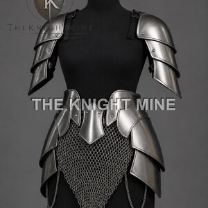 May include: Silver medieval armour set on a black mannequin. The armour includes shoulder pauldrons, a waist belt, and a chainmail skirt. The text "THE KNIGHT MINE" is visible on the armour.