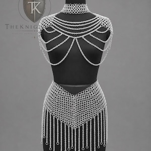 May include: A silver-toned chainmail body harness featuring a high-neck choker, draped shoulder chains, and a waist-length skirt with a V-shaped front and long dangling chain fringe. The piece is displayed on a black mannequin with a logo in the corner.