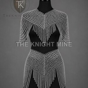 May include: A silver chainmail top and skirt set on a black mannequin. The chainmail is made of small, interlocking metal rings. The top has short sleeves and a fringed hem. The skirt has a fringed hem. The text "THE KNIGHT MINE" is visible.