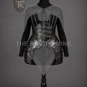 May include: A black long-sleeved garment worn under a dark metal corset with three horizontal leather straps and buckles. The ensemble features a silver chainmail shoulder mantle and a pointed chainmail skirt overlay, with the text THE KNIGHT MINE.