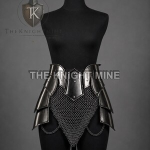 May include: A dark metal tasset belt featuring layered steel plates and a central chainmail section. The armour piece includes small metal rivets and thin hanging chains. The text THE KNIGHT MINE is visible on the front of the armour and in the logo.