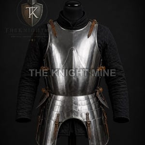 May include: A polished steel breastplate and tassets set featuring a central vertical ridge, brown leather straps with metal buckles, and a black quilted gambeson underneath. The text THE KNIGHT MINE is visible across the centre of the armour.