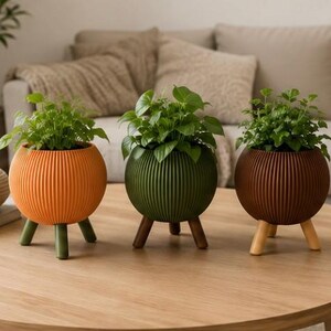 Pode incluir: Três vasos esféricos com plantas verdes sobre uma mesa de madeira. Os vasos são laranja, verde e castanho, cada um com uma textura canelada e três pernas de madeira. O fundo inclui um sofá bege e itens decorativos.