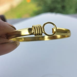 Peut inclure: Un bracelet jonc en métal doré avec une finition lisse et polie. Le design comprend un élément décoratif central composé d'une série de stries horizontales enroulées autour de l'anneau, relié à un mécanisme de fermoir en forme de cercle.
