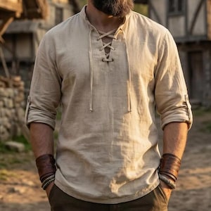 Peut inclure: Une chemise beige à manches longues dotée d'un laçage sur le devant avec des liens en tissu assortis au niveau de l'encolure. La chemise présente un col officier, des manches retroussées fixées par des pattes boutonnées et une finition texturée.