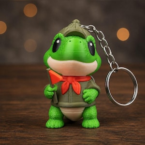 Könnte beinhalten: Ein grüner Frosch-Schlüsselanhänger, der eine Figur mit olivgrünem Pfadfinderhut und passender Uniform mit rotem Halstuch zeigt. Der Frosch hält eine kleine orangefarbene dreieckige Flagge in der Hand und hat eine silberne Metallkette mit Ring.