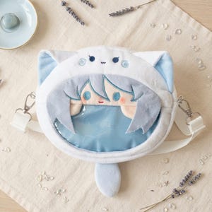 Peut inclure: Un sac bandoulière en peluche blanche en forme de tête de chat avec des oreilles bleu clair. L'avant présente une fenêtre transparente montrant un personnage brodé de style anime aux cheveux bleu clair, yeux bleus et une petite queue.