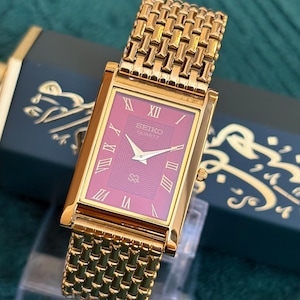 May include: A rectangular Seiko Quartz watch with a gold-tone metal link bracelet and a deep magenta dial. The watch face features gold-tone Roman numeral hour markers, gold-tone hands, and the Seiko logo printed in white at the top center.