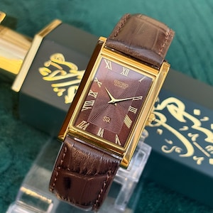 Könnte beinhalten: Eine rechteckige, goldfarbene Seiko Quartz Armbanduhr mit einem dunkelbraunen, strukturierten Zifferblatt, goldenen römischen Ziffern als Stundenmarkierungen und einem braunen Armband aus geprägtem Leder mit goldener Beschriftung.