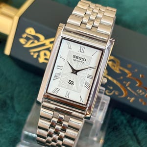 Puede incluir: Un reloj Seiko Quartz rectangular con una correa plateada y esfera blanca. La esfera del reloj presenta números romanos y agujas negras, con el logotipo de Seiko y un pequeño emblema. El reloj se exhibe sobre un soporte transparente.