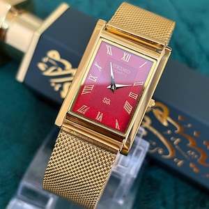 May include: A gold-toned rectangular Seiko Quartz watch with a red face and gold Roman numerals. The watch has a mesh band and is displayed on a clear stand. The watch is set against a dark green background.