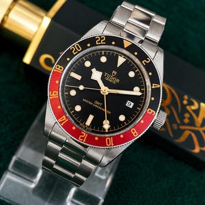 Puede incluir: Un reloj de pulsera Tudor GMT de acero inoxidable con esfera negra, marcadores de hora luminosos y un bisel giratorio bidireccional con un inserto rojo y negro con marcas de 24 horas. El reloj incluye una ventana de fecha y una pulsera de eslabones metálicos.