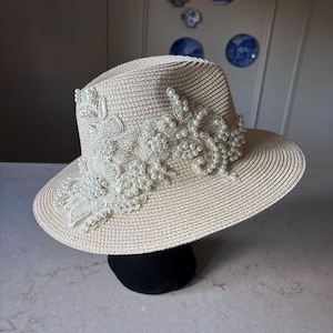 Op de afbeelding: Een beige strooien fedora-hoed met een ingewikkelde witte kanten applicatie aan de zijkant. Het kantontwerp is rijkelijk versierd met talloze kleine, ronde, witte kunstparels die een gestructureerd, bloemachtig patroon op de kroon vormen.