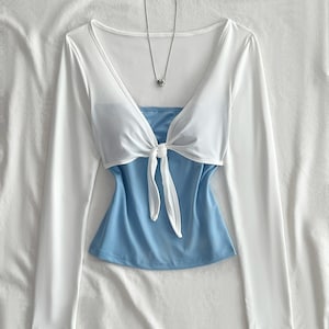 May include: A long-sleeved white top with a V-neckline and a front tie detail, layered over a solid light blue camisole. A thin silver-toned chain necklace with a small heart-shaped pendant is positioned at the neckline of the white top.