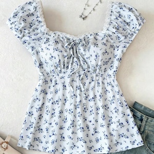 May include: A white short-sleeved top featuring a blue floral pattern, a square neckline with white lace trim, and a center front tie closure. The garment has puffed sleeves and a ribbed texture, displayed alongside a necklace and denim fabric.
