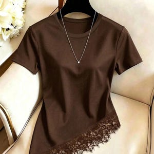 Puede incluir: Una camiseta de manga corta de color marrón oscuro con cuello redondo y un dobladillo asimétrico adornado con encaje marrón a juego. Un delicado collar de cadena plateada con un pequeño colgante circular descansa sobre la tela.
