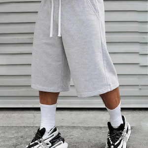 May include: Light gray fleece shorts featuring an elastic waistband with a white drawstring, two side pockets, and a loose, knee-length fit. The shorts are paired with white crew socks and black and white athletic sneakers with thick soles.