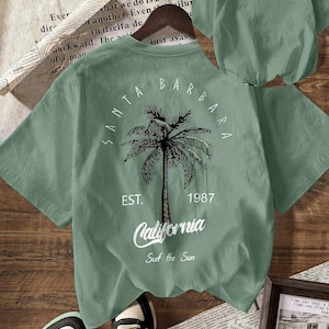 May include: A sage green short-sleeve t-shirt featuring a palm tree graphic on the back with the text Santa Barbara, Est. 1987, California, and Surf the Sun. The front chest area displays the word California in white script lettering.