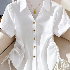 May include: A crisp white short-sleeved button-down shirt featuring a classic collar, five gold-toned circular buttons down the center, and adjustable drawstring ruching details on both sides of the waist with dangling white fabric ties.