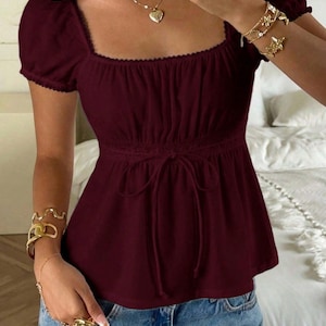 May include: A dark burgundy short-sleeved blouse featuring a square neckline, a gathered bodice, and a drawstring waist tie. The garment is paired with light blue denim jeans and accessorized with gold-toned necklaces and multiple gold-toned bracelets.