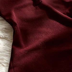 May include: A deep burgundy long-sleeve shirt featuring a vertical ribbed texture and a partial button-down placket at the neckline with three small, light-colored circular buttons. The fabric has a soft, fitted appearance with visible vertical lines.