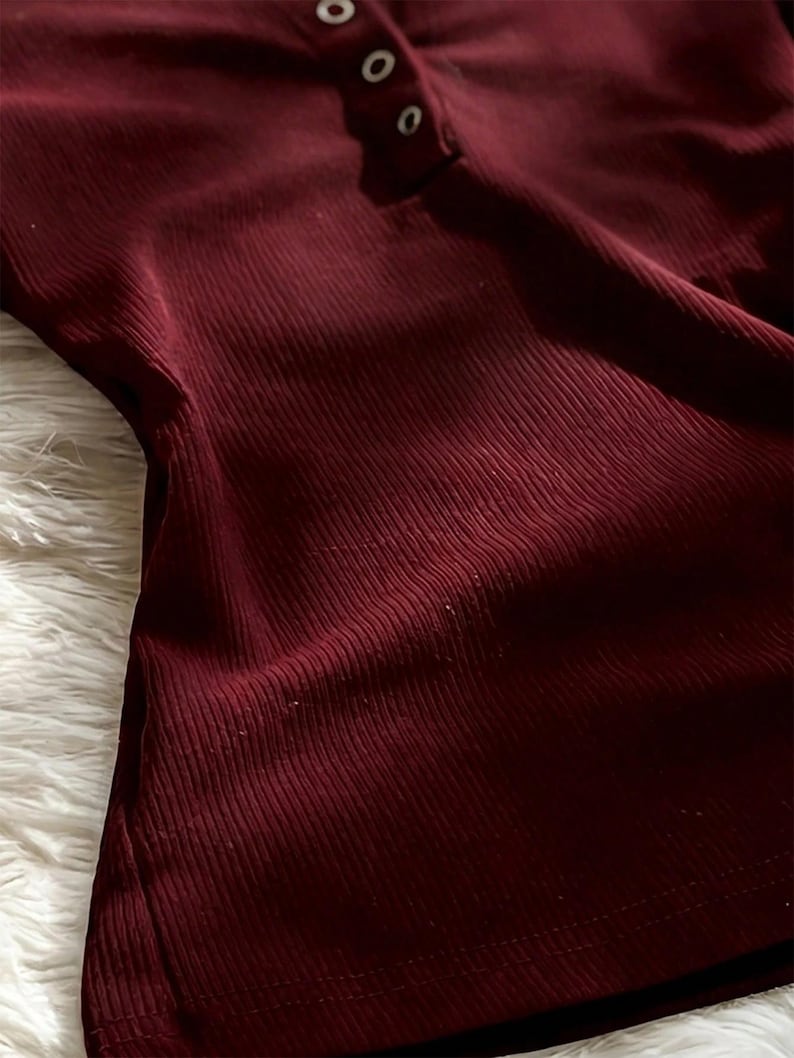May include: A deep burgundy long-sleeve shirt featuring a vertical ribbed texture and a partial button-down placket at the neckline with three small, light-colored circular buttons. The fabric has a soft, fitted appearance with visible vertical lines.