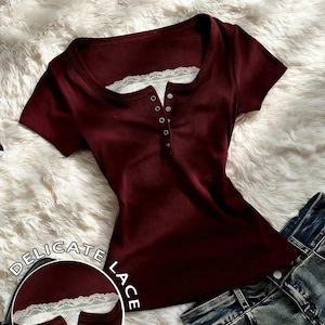 May include: A short-sleeved, ribbed, burgundy henley shirt featuring a square neckline accented with white lace trim and a front button placket. A circular inset shows a close-up of the lace detail with the text DELICATE LACE printed nearby.