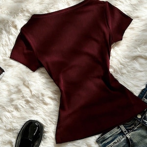 May include: A short-sleeved, ribbed burgundy t-shirt with a scoop neckline. The shirt is displayed alongside a pair of blue denim jeans and black patent leather shoes, showcasing a simple, solid-colored garment with a fitted silhouette.