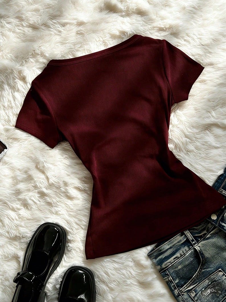 May include: A short-sleeved, ribbed burgundy t-shirt with a scoop neckline. The shirt is displayed alongside a pair of blue denim jeans and black patent leather shoes, showcasing a simple, solid-colored garment with a fitted silhouette.