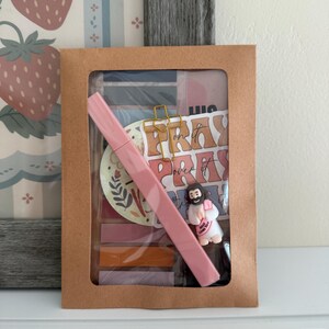 May include: A brown paper envelope with a clear window containing a pink highlighter, a gold cross-shaped paperclip, a small figurine of a person with a sign reading Jesus Loves You, and stickers with the text Pray On It Pray Over It.