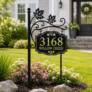 May include: A black metal address sign featuring the number 3168 and the text WILLOW CREEK. The sign is mounted on a vertical post with decorative scrollwork and two maple leaf silhouettes at the top, positioned in a garden bed.