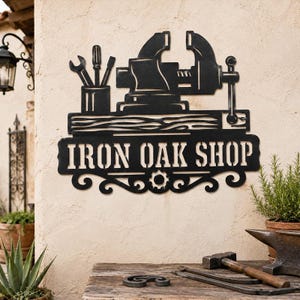May include: A black metal wall sign featuring a silhouette of a bench vise and tools in a holder above a textured horizontal bar. The text IRON OAK SHOP is cut out in capital letters, with decorative scrollwork and a gear icon at the bottom.