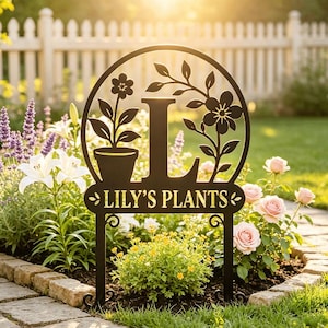 May include: A black metal garden sign featuring a circular frame with a cutout letter L, a potted plant, and a floral vine. The sign includes the text LILY'S PLANTS in gold lettering and is supported by two decorative metal stakes.