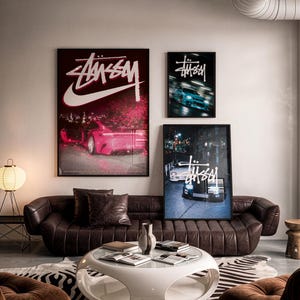 Puede incluir: Una sala de estar con un sofá capitoné marrón oscuro, una mesa de centro circular blanca y tres pósteres enmarcados en la pared. Los pósteres muestran el logotipo de Stüssy junto a imágenes de un coche deportivo rosa y otros dos vehículos.
