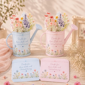 Könnte beinhalten: Zwei Gießkannen aus Metall in Hellblau und Hellrosa, gefüllt mit Blumenarrangements aus Papier. Beide Kannen und die dazugehörigen flachen Karten zeigen den Text Danke für deine Liebe und Unterstützung sowie einen bunten Wildblumenaufdruck.