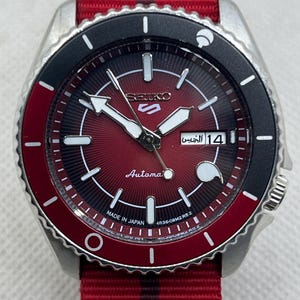 May include: A Seiko 5 automatic wristwatch with a red and black sunray dial, silver-tone hands, and indices. The watch features a day-date window, a black and red rotating bezel, and a red nylon strap with a central black stripe.