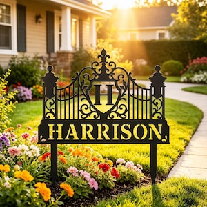 May include: A black metal garden sign featuring an ornate gate-style design with a central shield displaying the letter H and the name HARRISON in bold capital letters. The sign is supported by two stakes inserted into the ground.