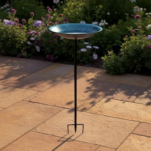 Può includere: Una vasca per uccelli turchese con una superficie strutturata su un supporto in metallo nero. Il supporto ha una base a tre punte. La vasca per uccelli è circondata da un giardino con fogliame verde e fiori viola e bianchi.