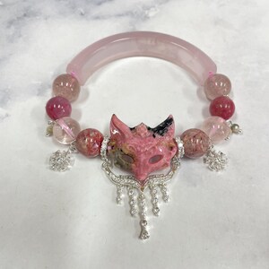 Puede incluir: Una pulsera con una barra curva de piedra rosa translúcida, cuentas redondas de piedra rosa y transparente, y un colgante central con forma de cabeza de zorro tallada. El zorro está decorado con metal plateado, cristales transparentes y dijes.