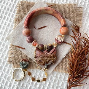 Puede incluir: Una pulsera con un gran segmento de piedra curvado de color marrón rojizo, cuentas redondas pulidas en tonos marrón y crema, y un colgante central con forma de cabeza de zorro tallada. Una cadena dorada conecta al zorro con un anillo separado.