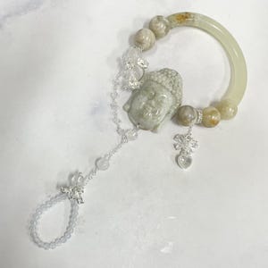 Puede incluir: Un adorno de mano con un brazalete verde pálido conectado mediante una cadena plateada a un anillo de cuentas más pequeño. La pieza incluye una cabeza de Buda tallada, cuentas moteadas, cristales transparentes y un dije en forma de corazón.