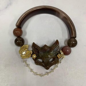Könnte beinhalten: Ein Armband mit einem gebogenen braunen Band, glatten braunen Kugeln und einem zentralen Anhänger in Form einer geschnitzten Fuchsmaske. Das Design umfasst ein goldfarbenes Ginkgo-Blatt-Element, kleine Blumenanhänger und eine feine Kette.