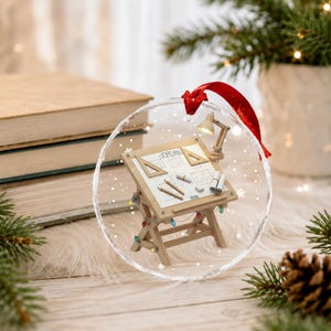 May include: A round clear glass ornament featuring a miniature wooden drafting table with the name JORDAN and the year 2026 printed on the surface. The table includes tiny drafting tools, a lamp, and is decorated with small colorful lights.