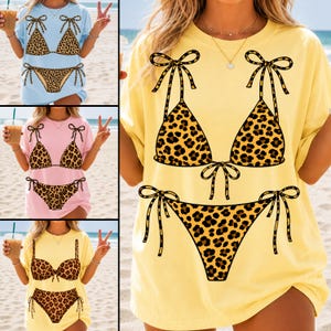 May include: A light blue, pink, and yellow t-shirt with a leopard print bikini design. The bikini top and bottom are brown with black spots and tied with black bows. The shirts are displayed on a beach.