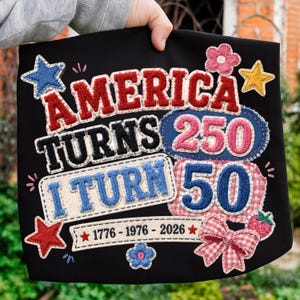 Peut inclure: Un article en tissu noir avec des écussons brodés portant le texte AMERICA TURNS 250 I TURN 50 et 1776-1976-2026. Le motif comprend des étoiles rouges, blanches et bleues, une fleur rose, une fraise rouge, un nœud vichy rose et des lettres texturées.