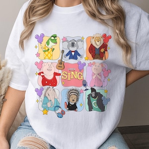 May include: A white t-shirt featuring a graphic print of characters from the movie Sing, including a koala, lion, pig, elephant, gorilla, and porcupine. The design includes colorful rounded squares, balloons, a guitar, and the text Sing.
