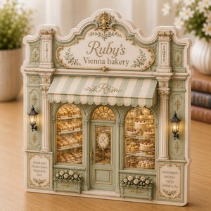 May include: A decorative miniature facade of Ruby's Vienna bakery in sage green and cream. The structure features a striped awning, two illuminated black wall lamps, and display windows filled with miniature bread, cakes, and pastries.